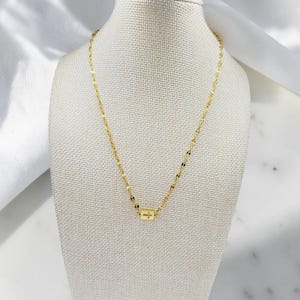 Puede incluir: Un collar de tono dorado con una cadena delicada de eslabones planos y reflectantes, que presenta un pequeño colgante rectangular centrado. El colgante muestra un diminuto símbolo de cruz grabado en su superficie frontal, creando un diseño sutil.