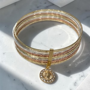 May include: A tri-color bangle bracelet featuring alternating bands of gold, silver, and rose gold tones with a textured, star-like pattern. A circular gold-toned pendant with a central figure and a border of small clear stones hangs from the bangle.