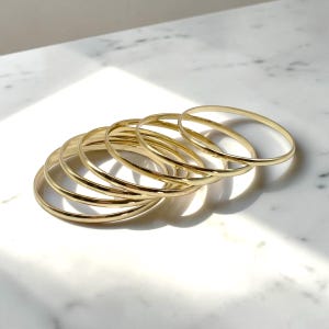 May include: A set of seven thin, polished gold-tone metal bangles arranged in a slightly overlapping, cascading formation. Each circular bracelet features a smooth, reflective surface and a simple, continuous band design with no additional embellishments.