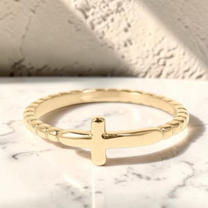 May include: A gold-toned ring featuring a smooth, polished cross centerpiece integrated into a textured, beaded band. The ring has a reflective metallic finish and a circular shape, showcasing a simple design with a cross motif as the focal point.