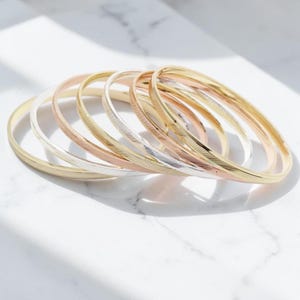 Può includere: Un set di sette braccialetti rigidi sottili e circolari in tonalità metalliche alternate di oro, oro rosa e argento. Ogni braccialetto presenta una sottile trama incisa in diagonale sulla superficie, creando un delicato motivo riflettente.