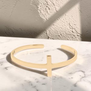 May include: A gold-toned open cuff bracelet featuring a centered cross design. The interior of the band displays the engraved text 4:13. The bracelet has a smooth, polished finish and a circular shape with a small opening at the back.