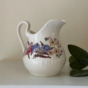 May include: A cream-colored ceramic pitcher featuring a scalloped rim, a curved handle, and a colorful floral pattern with blue, yellow, and red blossoms. The pitcher has a rounded body and a smooth, glossy finish, showcasing detailed botanical illustrations.