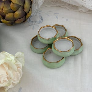 May include: A set of six small, round ceramic dishes with a pale green exterior and a white interior. Each dish features a scalloped edge rimmed with a thin line of gold paint. The dishes are arranged in a cluster, showcasing their uniform design.