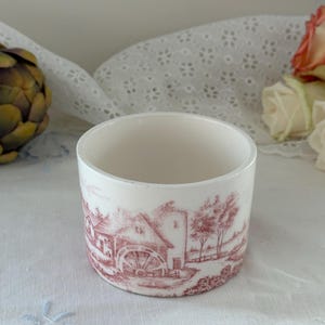 May include: A white ceramic cup featuring a detailed red transferware print of a rural landscape with a water mill. The cylindrical vessel has a smooth finish and a simple rim, showcasing a pastoral scene with trees and buildings in a monochromatic red ink.