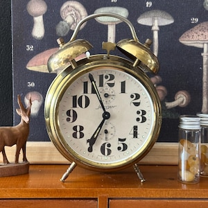 May include: A vintage brass Jaeger alarm clock with a cream-colored face and black numerals. The clock has a classic design with two bells on top and stands on four small legs. A wooden deer figurine and a mushroom print are in the background.