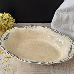May include: An oval ceramic serving dish with a cream-colored crackle glaze finish. The scalloped rim features a dark, intricate floral pattern along the edges, providing a decorative contrast to the smooth, aged appearance of the central basin.