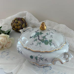 May include: A white porcelain tureen with a lid featuring a gold-colored finial. The tureen is decorated with green floral patterns and gold trim along the edges. It has two side handles and a scalloped base, showcasing a delicate botanical design.