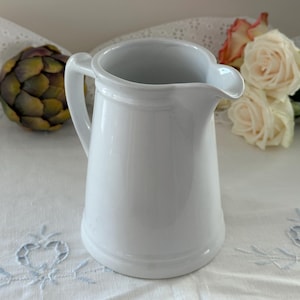 May include: A plain white ceramic pitcher with a smooth, glossy finish, a curved handle on the side, and a small pouring spout at the top rim. The pitcher has a cylindrical body that tapers slightly toward the top and a rounded base.