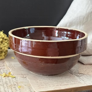 May include: A round, deep ceramic mixing bowl with a glossy, dark brown glaze. The bowl features a distinct, unglazed cream-colored band around the upper rim and a secondary horizontal ridge encircling the middle of the exterior body.