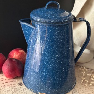 May include: A blue enamelware coffee pot with a lid and handle. The pot has a speckled pattern of white dots on a blue background. The pot is sitting next to three red peaches.