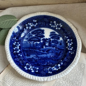 May include: A round ceramic plate featuring a deep blue and white transferware pattern. The center depicts a landscape with a stone bridge, trees, and buildings. The scalloped rim is decorated with a floral motif in matching blue and white.