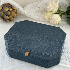 May include: A dark blue, octagonal jewelry box with a textured, pebbled surface finish. The box features a small, centered gold-toned metal knob on the front edge for opening. The lid is flat and the sides are angled to create an eight-sided shape.