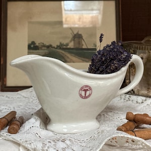 May include: A white ceramic gravy boat featuring a small circular red emblem with a caduceus symbol and the text United States Army Medical Department. The vessel is filled with a cluster of dried purple lavender sprigs and has a curved handle.