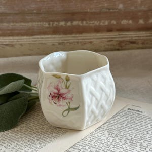 May include: A small, off-white ceramic cup with a scalloped rim and a textured basket-weave pattern on its exterior. The cup features a delicate, hand-painted illustration of a pink lily flower with green leaves on one side of its curved body.