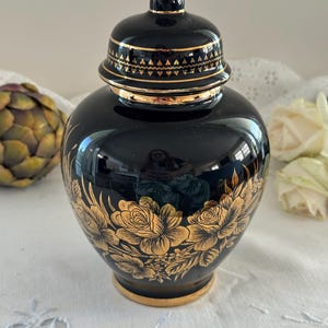 May include: A black ginger jar with a matching lid, featuring a gold floral pattern around the base and a gold geometric band on the lid. The jar has a glossy finish, a rounded body, and a gold-trimmed base, reflecting light on its surface.