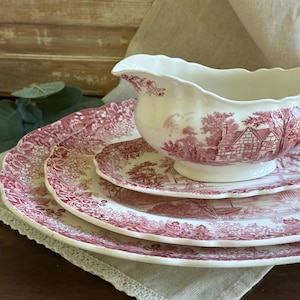 May include: A cream-colored ceramic gravy boat and two matching plates featuring a red transferware pattern of a rural cottage landscape with trees and floral borders. The pieces have scalloped edges and a glossy finish.
