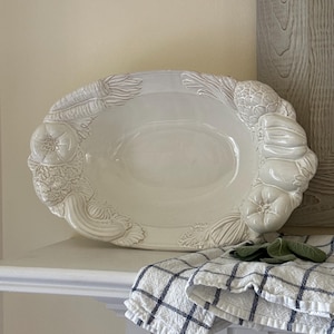 May include: An oval-shaped white ceramic serving platter featuring a raised, embossed relief pattern of various vegetables, including carrots, peppers, and artichokes, along the outer rim. The platter has a glossy finish and a smooth, concave center.