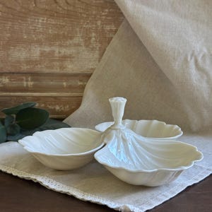 May include: A white milk glass serving dish featuring three shell-shaped bowls arranged around a central vertical handle. The piece has a glossy finish and scalloped edges, with a molded design that mimics the appearance of natural seashells.