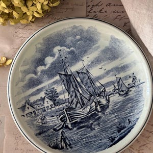May include: A round ceramic plate featuring a blue and white transferware print of a nautical scene. The illustration depicts two sailboats on a body of water, with a small village, a windmill, and a cloudy sky in the background. The plate has a thin dark blue rim.