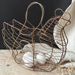 May include: A vintage-style wire egg basket shaped like a hen, with a handle and a head. The basket is made of copper-colored metal and contains three white eggs. The basket sits on a table with vintage paper.