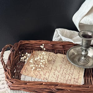 May include: A brown wicker tray holds a vintage letter, dried flowers, and a silver candlestick holder. The letter has handwritten text. A white cloth is in the background.