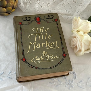 May include: A vintage hardcover book titled The Title Market by Emily Post with a muted olive green cover. The front features gold embossed lettering, two black crown illustrations, and a decorative border with small red oval accents.