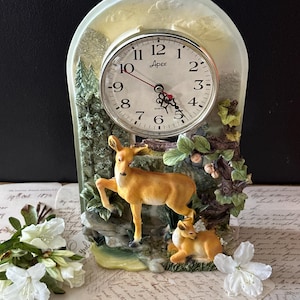 May include: A decorative clock featuring a deer family scene. The clock face is round with black numerals and hands, set against a backdrop of a forest scene with two deer. The clock is approximately 6 inches tall.