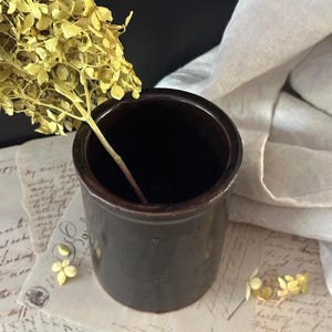 May include: A dark brown cylindrical ceramic crock with a glossy finish, containing a single stem of dried yellow hydrangea flowers. The crock is positioned on top of several pieces of vintage paper featuring cursive handwriting and postal stamps.