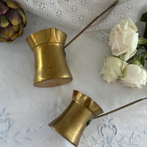 May include: Two brass coffee pots with long, thin handles and flared rims. The pots have a smooth, metallic finish with a warm, golden hue. Each vessel features a wide base that tapers inward toward the center before widening at the top opening.