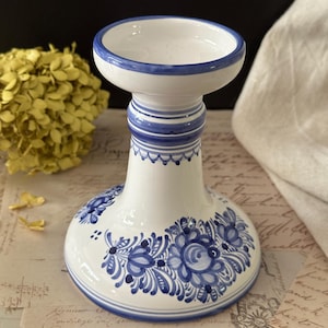 May include: A white ceramic candle holder with a flared base and a cylindrical top. The piece features hand-painted blue floral motifs around the base and horizontal blue bands around the neck and rim, creating a decorative contrast on the surface.