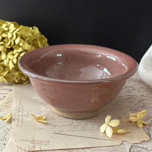 May include: A small, round ceramic bowl with a glossy, dusty rose-colored glaze. The bowl features a smooth, curved rim and a thick, unglazed beige base. The exterior surface has subtle, faint floral impressions embossed into the clay.