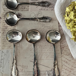 May include: Five silver-toned metal spoons with ornate, embossed handles featuring intricate scrollwork and engraved monograms. The spoons have round bowls and polished surfaces that reflect the surrounding environment, arranged in a group.