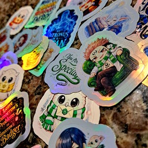 May include: A collection of holographic stickers with various designs. Some stickers feature anime-style characters, owls, and text like "My Patronus is a Fan" and "You Are Special". The stickers have a shiny, iridescent finish.