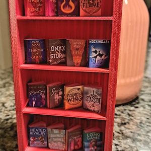 May include: A red miniature bookshelf filled with tiny book replicas. The books have colorful covers and titles like "A Court of Thorns and Roses" and "Mockingjay". A decorative item for book lovers.