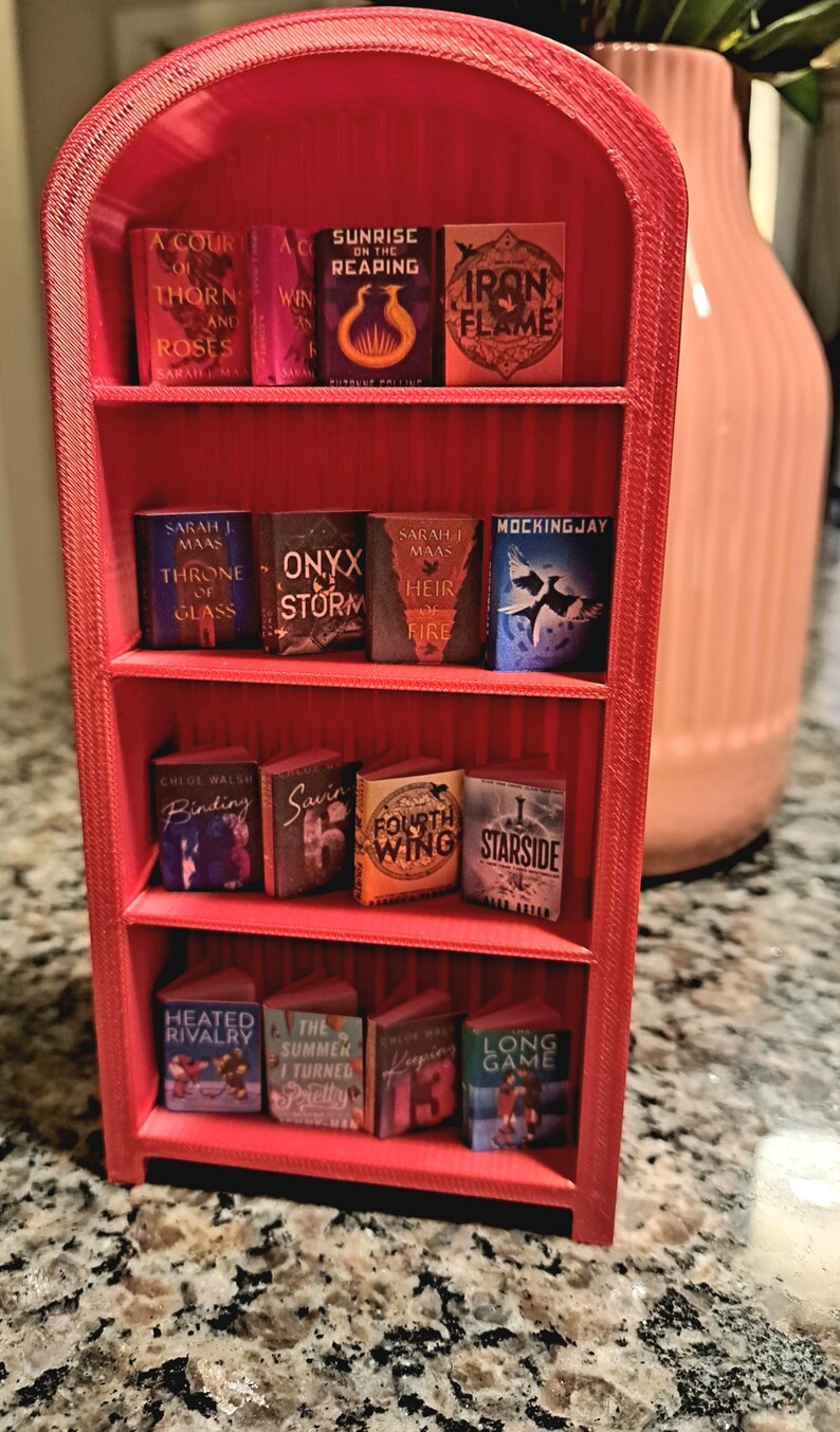 May include: A red miniature bookshelf filled with tiny book replicas. The books have colorful covers and titles like "A Court of Thorns and Roses" and "Mockingjay". A decorative item for book lovers.