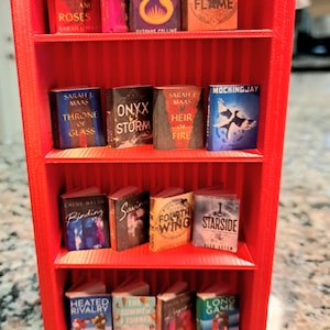 May include: A miniature red bookshelf filled with tiny books. The books have colorful covers and titles like "A Court of Thorns and Roses," "Sunrise on the Reaping," and "Iron Flame." The bookshelf is made of red plastic and has multiple shelves.