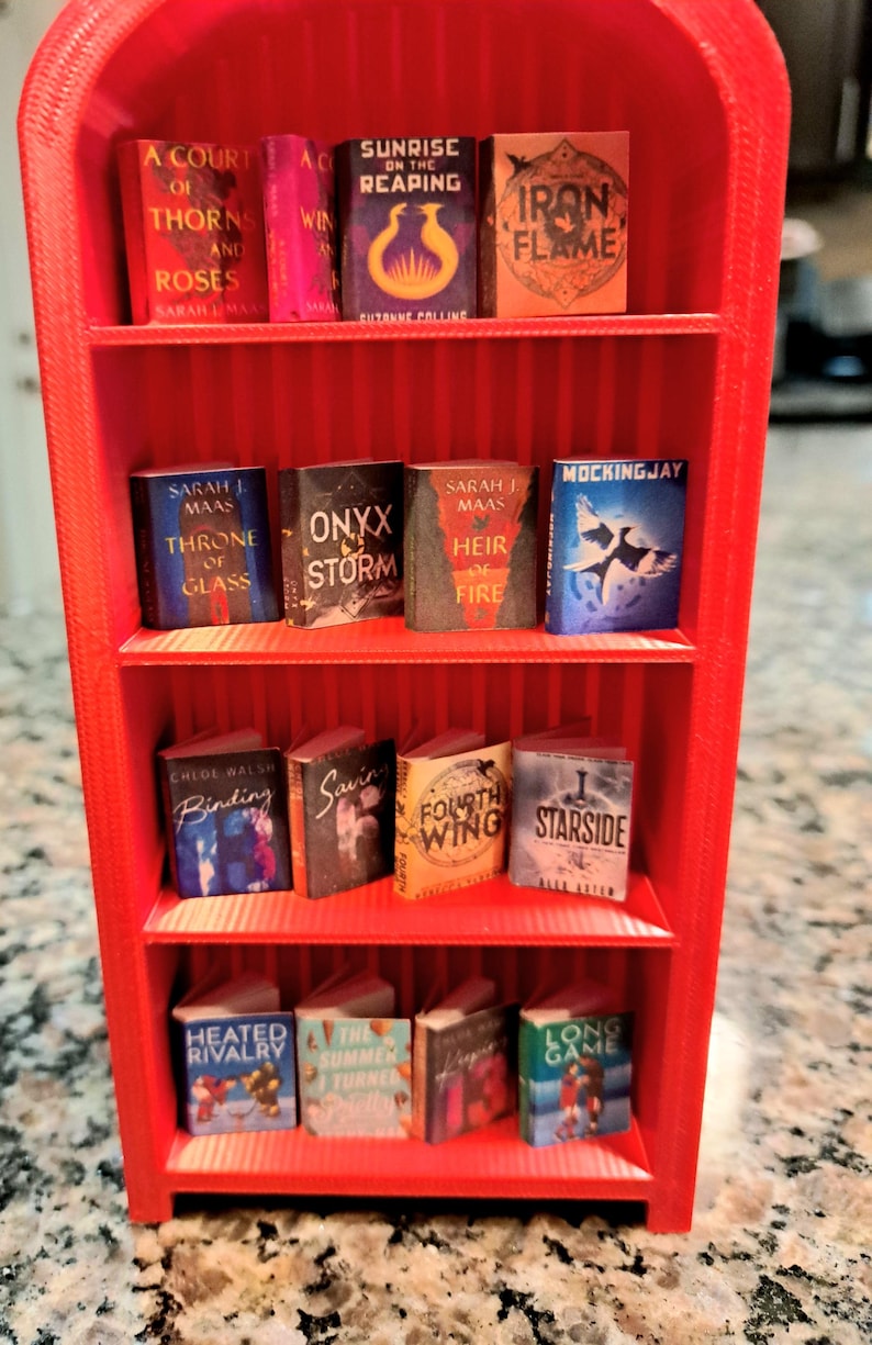 May include: A miniature red bookshelf filled with tiny books. The books have colorful covers and titles like "A Court of Thorns and Roses," "Sunrise on the Reaping," and "Iron Flame." The bookshelf is made of red plastic and has multiple shelves.