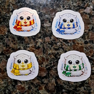 以下が含まれることがあります： 大きな目とふわふわの白い羽を持つ漫画のフクロウの4つのステッカー。各フクロウは、赤と金、青と金、黄と金、緑と白の異なる色のスカーフを着用しています。各フクロウは小さな封筒を持っています。