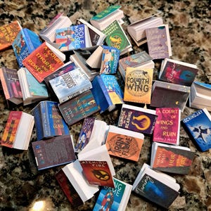 May include: A collection of miniature books with colorful covers and titles. The books are stacked and scattered, featuring titles like "A Court of Thorns and Roses," "Catching Fire," and "Fourth Wing." The books vary in size and color.