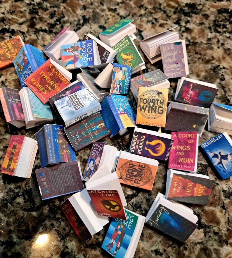 May include: A collection of miniature books with colorful covers and titles. The books are stacked and scattered, featuring titles like "A Court of Thorns and Roses," "Catching Fire," and "Fourth Wing." The books vary in size and color.