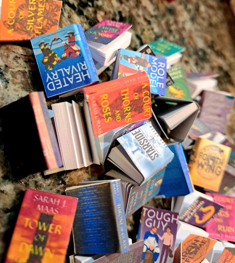 May include: A collection of miniature books with colorful covers and titles like "Heated Rivalry," "A Court of Thorns and Roses," and "Tower of Dawn." The books are stacked and scattered, showcasing various book cover designs and author names.