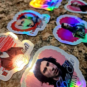 May include: Holographic stickers with various images and text. One sticker features a person with the text "I want a bad guy". Another says "If Vill Why Hot?". A third sticker says "I can fix him". The stickers have a pink and red border.