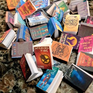 May include: A collection of miniature books with various titles and colorful covers. Titles include "A Court of Thorns and Roses," "Catching Fire," and "Fourth Wing." The books are arranged on a speckled surface, showcasing a variety of sizes and designs.