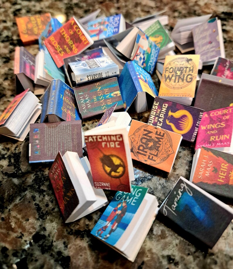 May include: A collection of miniature books with various titles and colorful covers. Titles include "A Court of Thorns and Roses," "Catching Fire," and "Fourth Wing." The books are arranged on a speckled surface, showcasing a variety of sizes and designs.