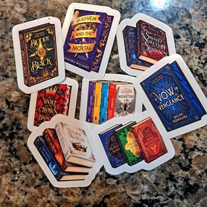 May include: A collection of rectangular vinyl stickers featuring various book cover illustrations. Titles include Bloody Black, Mayhem and the Mortal, Stalked by Seduction and Shadow, A Vow in Vengeance, The Wolf and the Crown, City of Gods and Monsters, City of Ruin, Wolf Witch, Heaven and Body, Fallen Star, and Holy Monsters.
