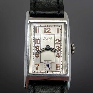 Puede incluir: Un reloj de pulsera rectangular Rolex Marconi con caja plateada y correa de cuero negro. La esfera blanca presenta números arábigos marrones, una subesfera de segundos en la parte inferior y manecillas negras. El texto Rolex Marconi está impreso.