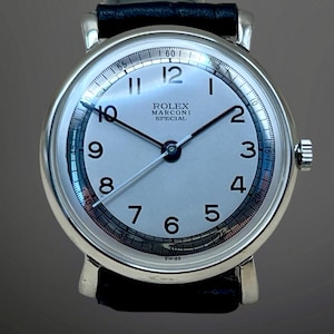 Könnte beinhalten: Eine silberfarbene Armbanduhr mit rundem weißem Zifferblatt, schwarzen arabischen Ziffern und dünnen schwarzen Zeigern. Das Zifferblatt trägt die Aufschrift ROLEX MARCONI SPECIAL und unten SWISS. Sie hat ein schwarzes Lederarmband und eine Krone.