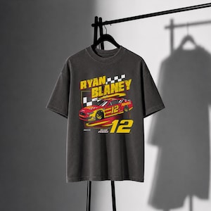May include: A charcoal gray short-sleeve t-shirt featuring a graphic print of a red and yellow race car with the number 12. The text reads RYAN BLANEY, 12, and TEAM PENSKE, accompanied by a checkered flag pattern and the NASCAR logo.