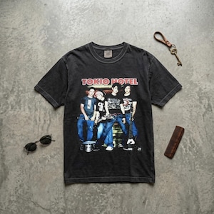 May include: A black Tokio Hotel band t-shirt featuring a graphic of the band. The shirt has a crew neck and short sleeves. Sunglasses, a key, and a comb are also visible.